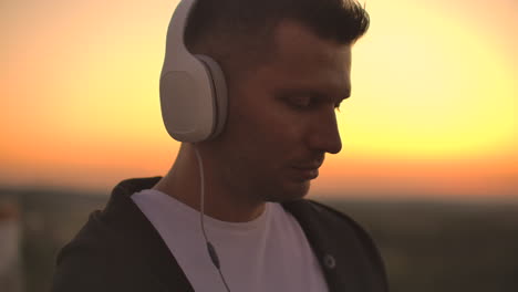 Smiling-and-laughing-handsome-young-man-listening-music-from-his-smartphone-in-wireless-headphones.-In-slow-motion-a-man-stands-on-the-roof-at-sunset-and-looks-at-the-beautiful-view-from-the-height-of-the-city